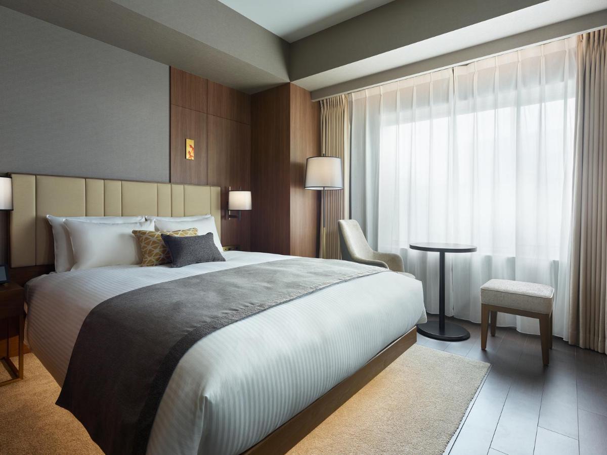 Mitsui Garden Hotel Roppongi Tokyo Premier Εξωτερικό φωτογραφία The photo shows a modern hotel room featuring a comfortable bed with white linens and a decorative throw pillow. The room has a neutral color palette with wooden accents on the walls. There are two bedside lamps and a chair positioned next to a small