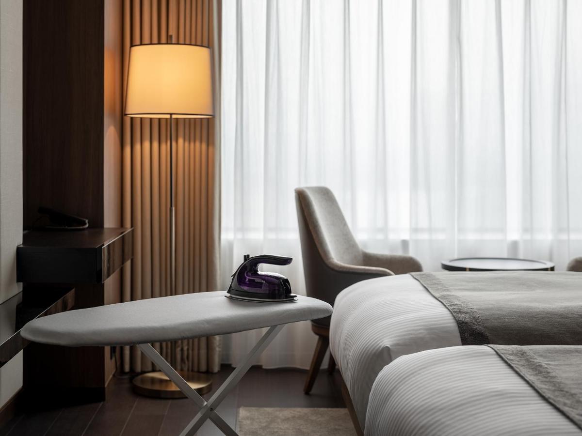 Mitsui Garden Hotel Roppongi Tokyo Premier Εξωτερικό φωτογραφία The photo shows a modern hotel room featuring two beds with white bedding. There is a light-colored armchair next to a small side table. On the table, there is an iron, and beside it, a lamp with a warm glow is illuminating the space. The large windo