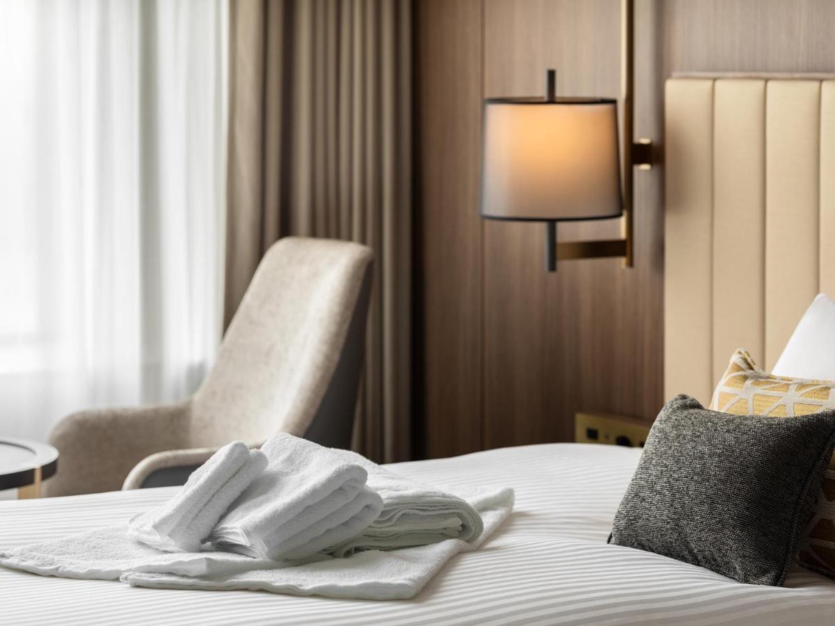 Mitsui Garden Hotel Roppongi Tokyo Premier Εξωτερικό φωτογραφία The image shows a stylish hotel room. In the foreground, there is a neatly made bed with white linens, featuring a stack of white towels arranged in a decorative manner. To the side, there is a gray upholstered armchair and a small table. The backgro