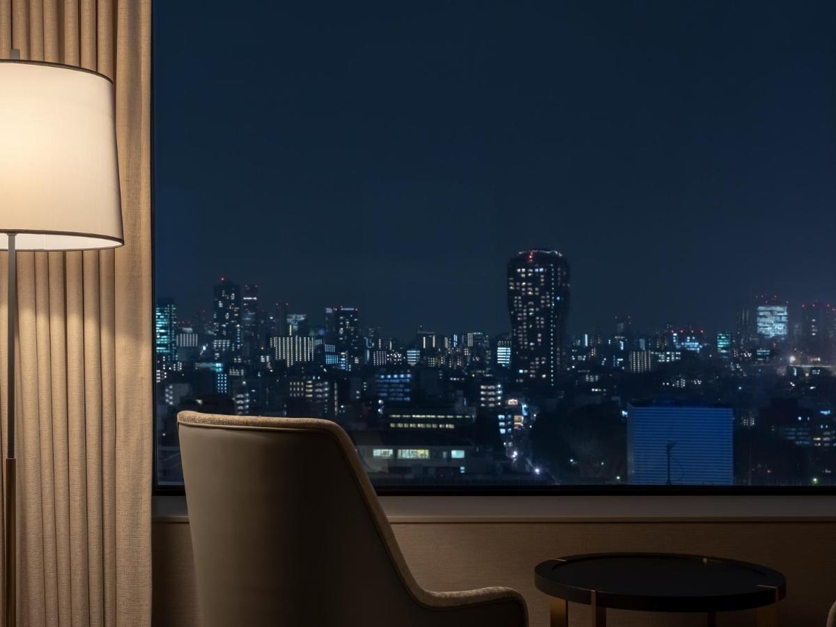 Mitsui Garden Hotel Roppongi Tokyo Premier Εξωτερικό φωτογραφία The photo depicts a cozy hotel room setting at night. In the foreground, there's a stylish armchair next to a small round table. A standing lamp provides soft lighting, creating a warm ambiance. In the background, large windows reveal a city skyline 