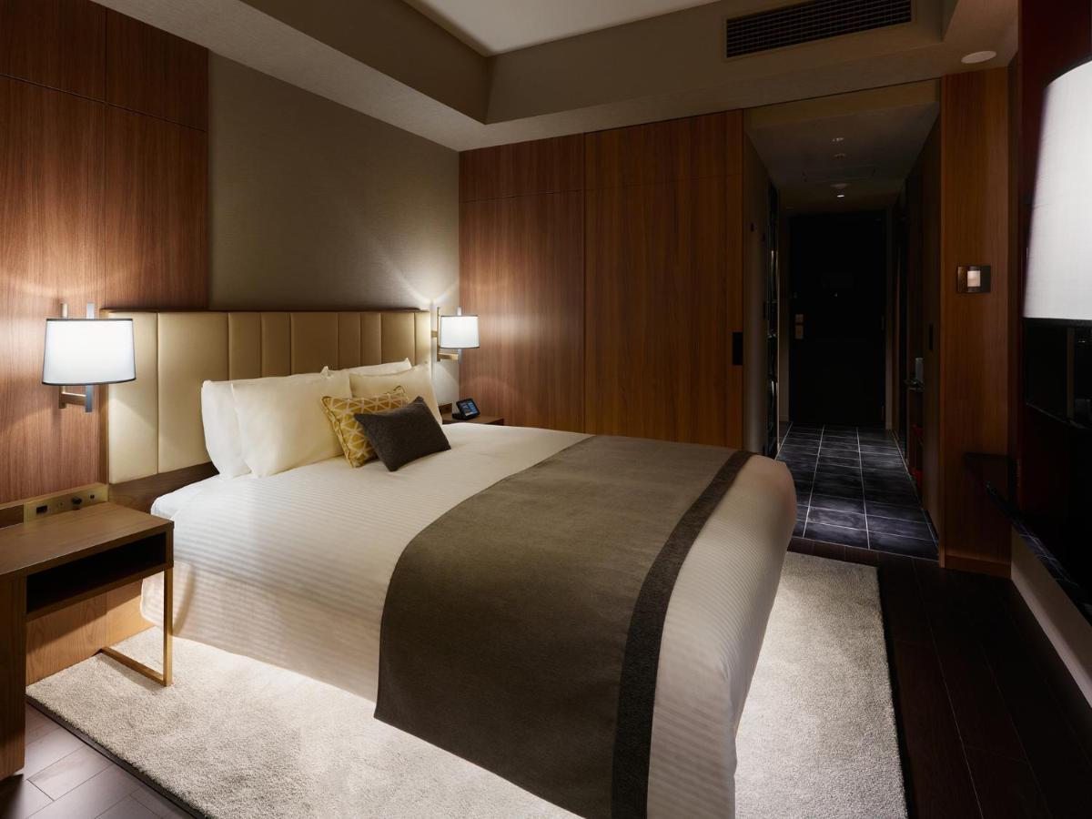 Mitsui Garden Hotel Roppongi Tokyo Premier Εξωτερικό φωτογραφία The image shows a modern hotel room with a warm and elegant design. In the center, there is a neatly made bed with white linens and a dark gray throw blanket. Flanking the bed are two bedside lamps providing soft lighting. The walls have a rich wood 