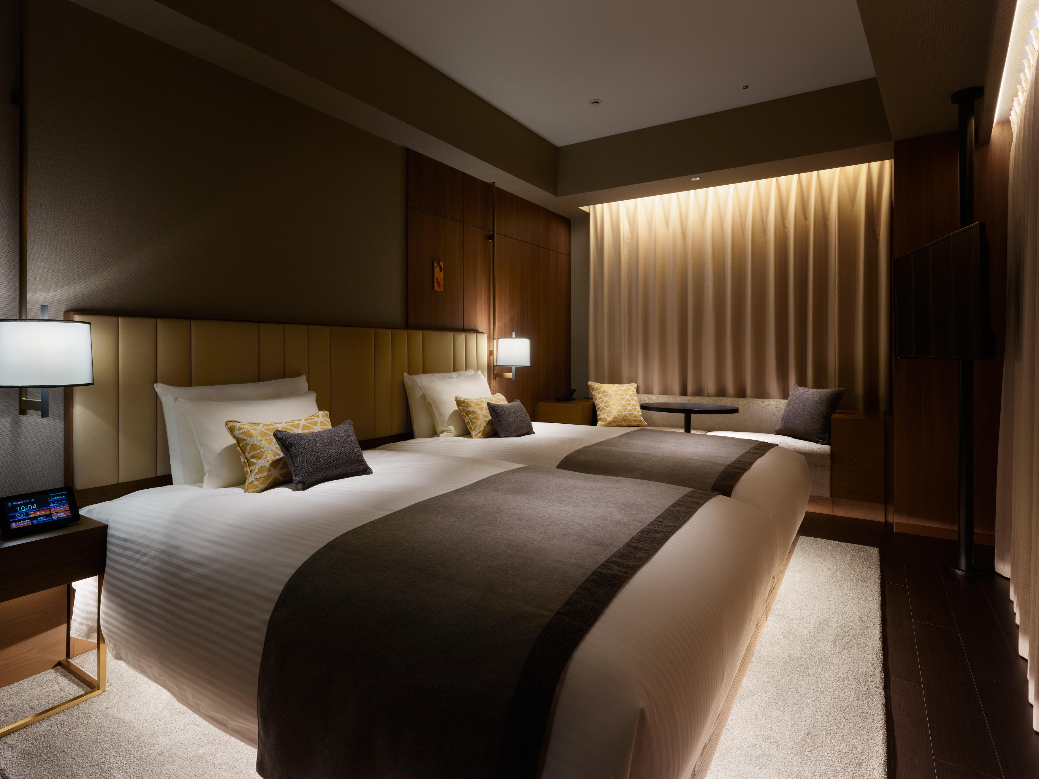 Mitsui Garden Hotel Roppongi Tokyo Premier Εξωτερικό φωτογραφία The photo shows a modern hotel room featuring two beds with plush white linens and decorative pillows. The room has a warm and inviting ambiance, highlighted by soft lighting from lamps and the backlit curtains. There is also a small table with chair