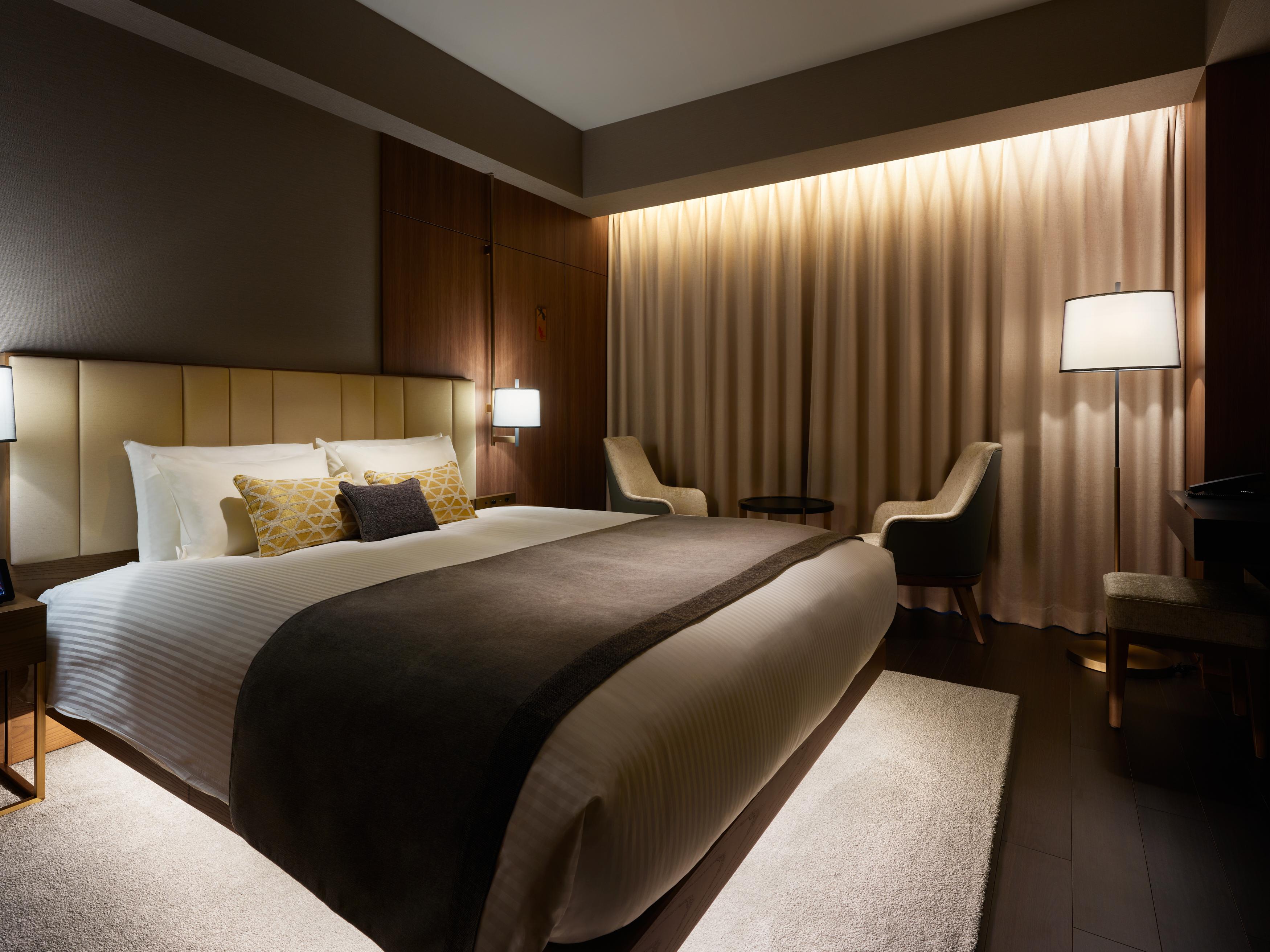 Mitsui Garden Hotel Roppongi Tokyo Premier Εξωτερικό φωτογραφία The photo depicts a modern hotel bedroom. It features a large bed with white linens and a dark gray throw blanket, accented by decorative pillows. The room has a warm ambiance created by soft lighting, including two bedside lamps and a lighted area b