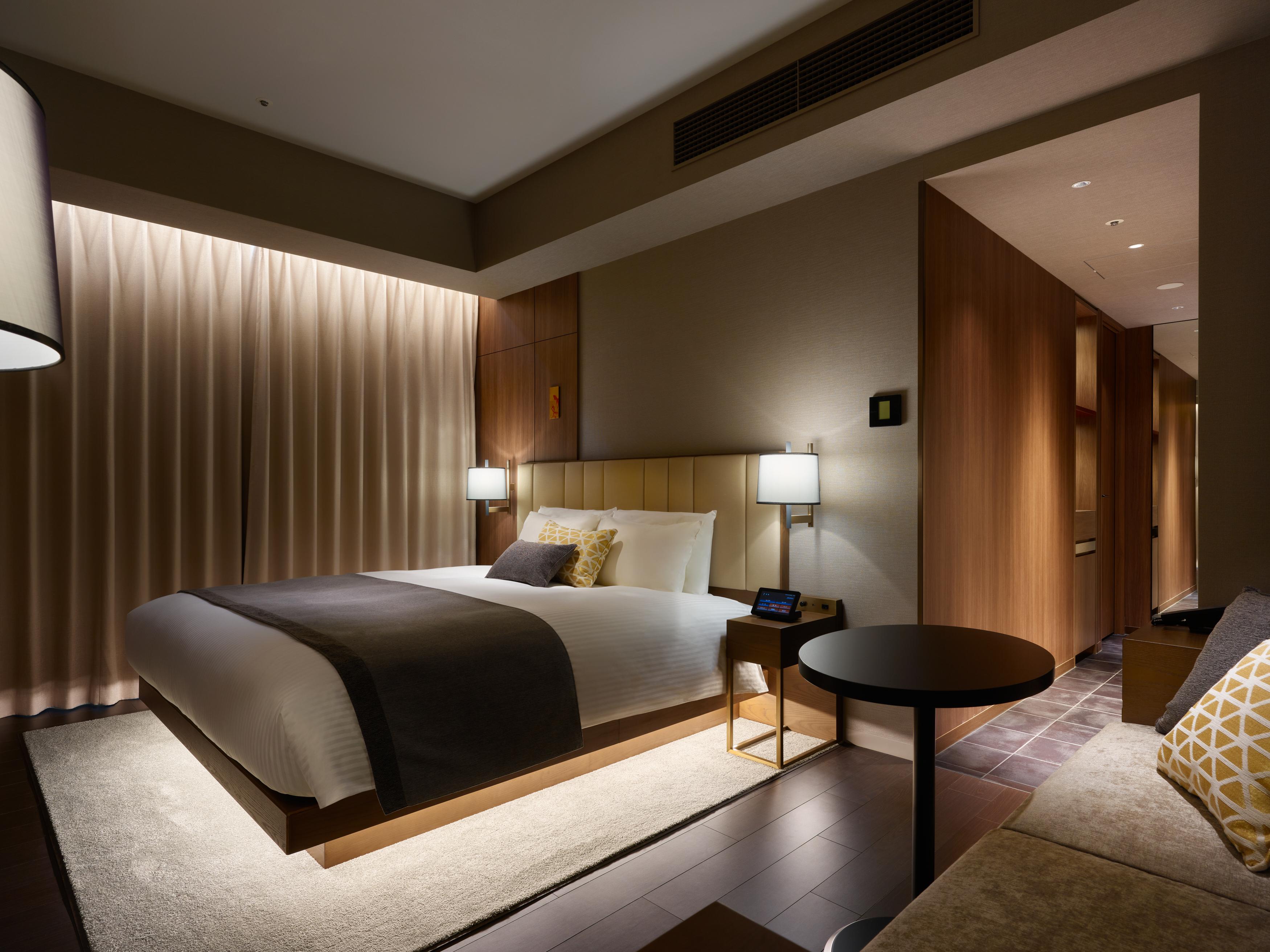 Mitsui Garden Hotel Roppongi Tokyo Premier Εξωτερικό φωτογραφία The photo shows a modern hotel room featuring a king-sized bed with neutral-colored bedding and accent pillows. There are two bedside lamps on either side of the bed, providing a warm glow. The walls have a wood finish, and there's a soft area rug on