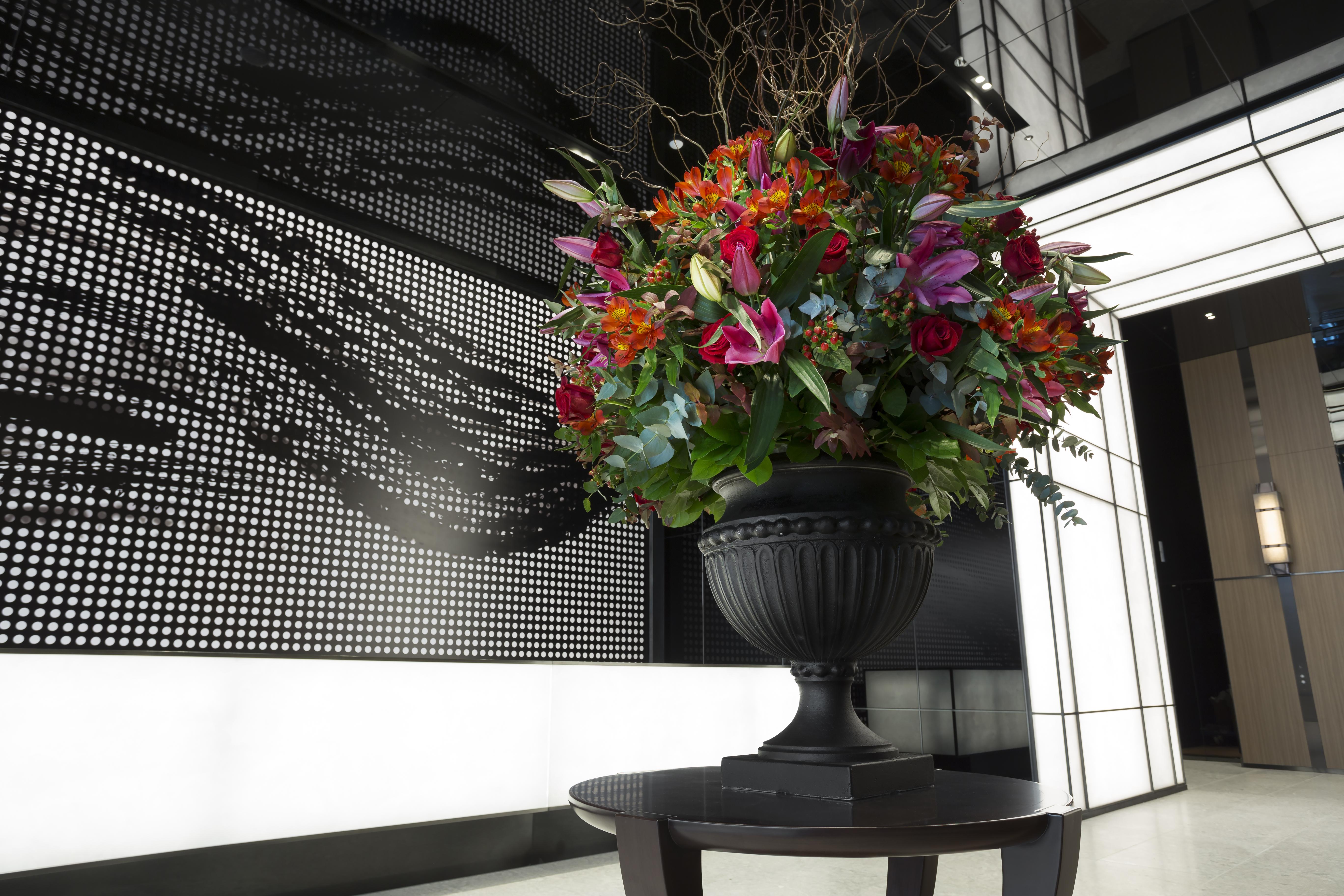 Mitsui Garden Hotel Roppongi Tokyo Premier Εξωτερικό φωτογραφία The photo features a large, vibrant floral arrangement in an ornate black urn. The arrangement is composed of various colorful flowers and greenery, showcasing shades of red, pink, orange, and green. This floral display is positioned on a small table