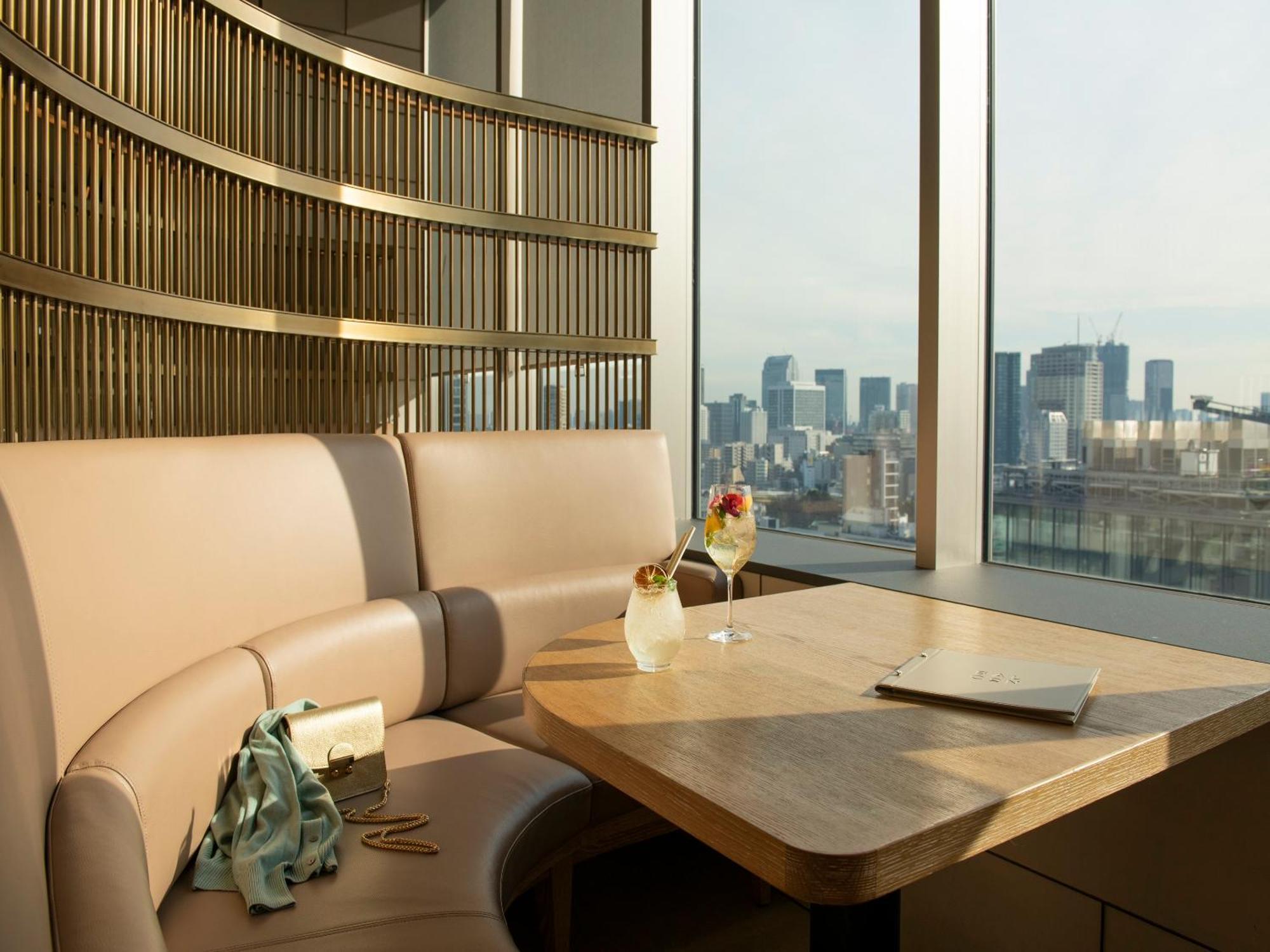 Mitsui Garden Hotel Roppongi Tokyo Premier Εξωτερικό φωτογραφία The photo depicts a cozy, modern seating area with a curved booth that features soft, pale upholstery. A wooden table is positioned in front of the booth, and on the table, there are two elegant drinks garnished with fruit. The setting has large wind