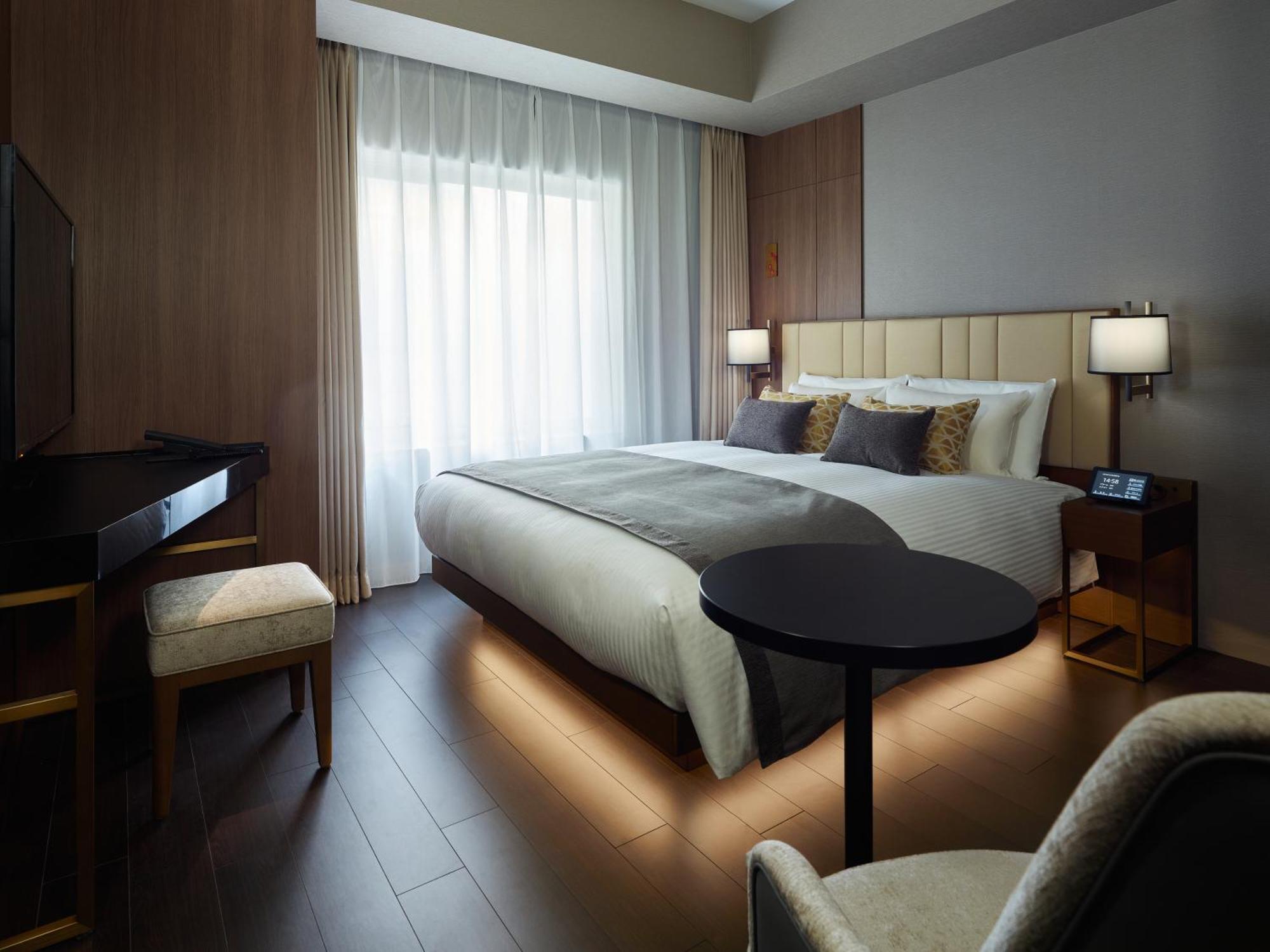 Mitsui Garden Hotel Roppongi Tokyo Premier Εξωτερικό φωτογραφία The photo shows a modern hotel room featuring a large bed with decorative pillows. There are large windows with sheer curtains allowing natural light to filter in. To one side, there is a sleek desk with a chair and a small round table in the center 